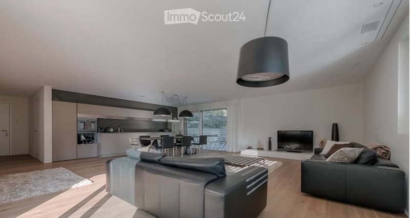 Modern, open-plan living and dining area with light wood flooring, white walls, and large windows. Features a grey leather sofa, coffee table, dining table with chairs, and a sleek kitchen area with cabinets and appliances.