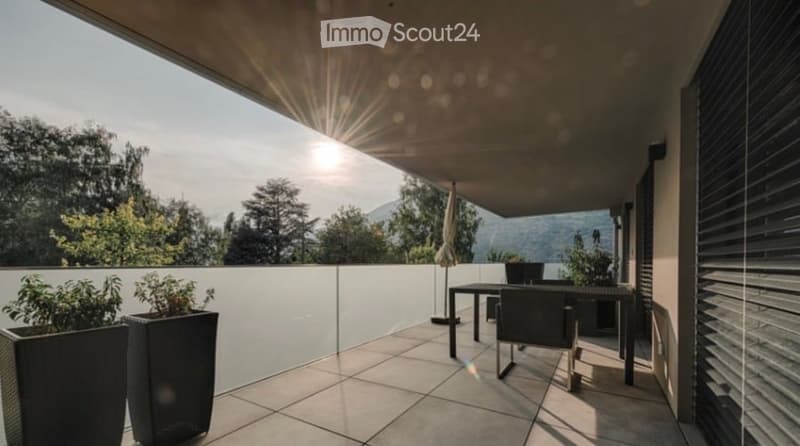 A modern balcony with a tiled floor, white glass railing, and outdoor furniture including a table and chairs. The balcony features potted plants and offers a scenic view of trees and mountains.