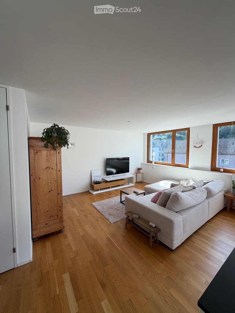 Modern living room with wooden flooring, large windows, a grey sofa, coffee table, TV, and decorative plants.
