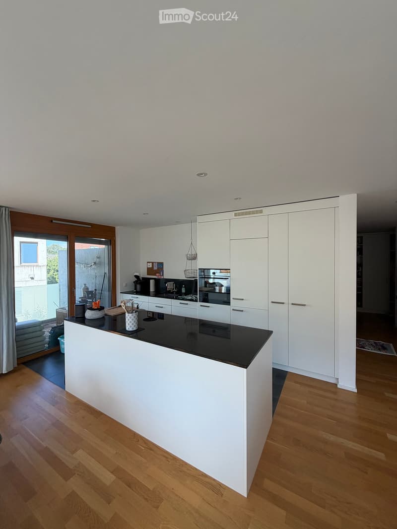 Modern kitchen with white cabinets, black countertop, wooden floor, and a large island. Features a sink, coffee maker, microwave, and various kitchen items on the counter.