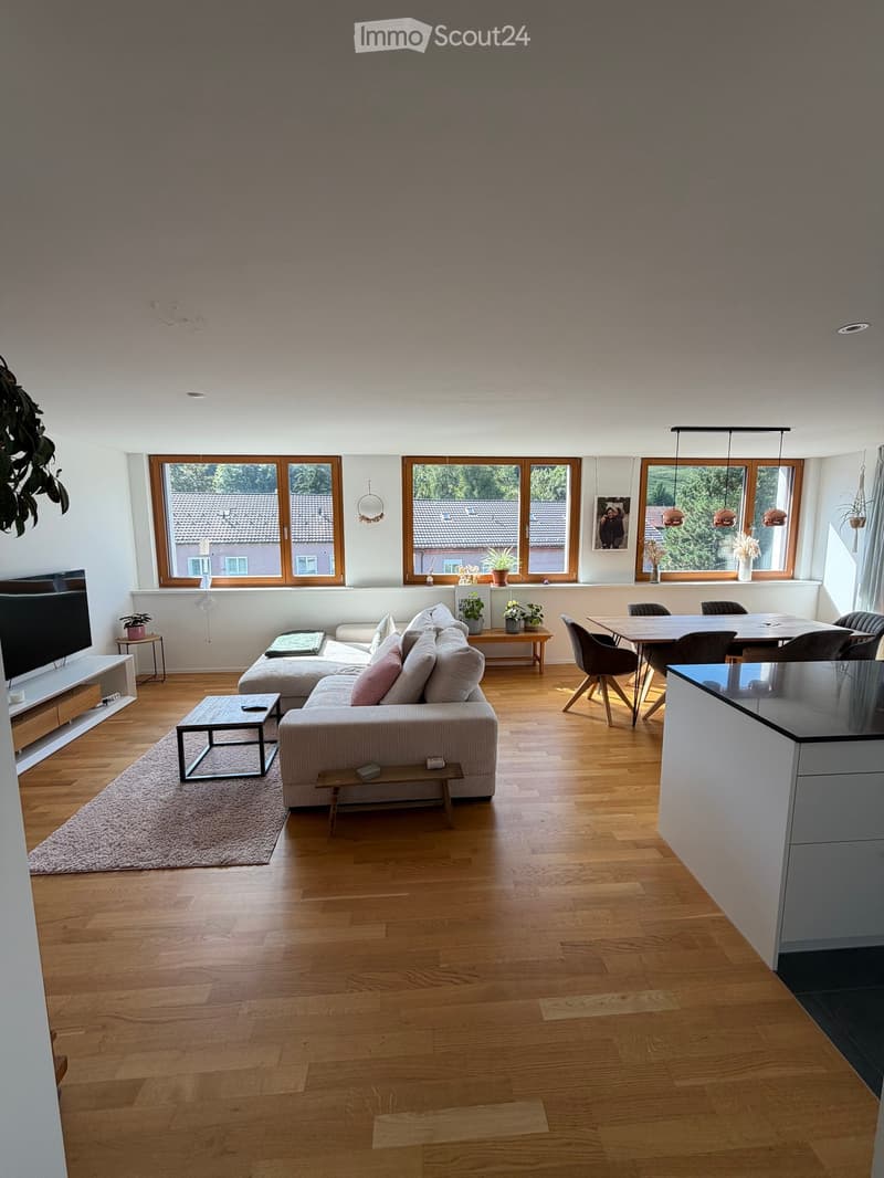 Open-plan living and dining area with wooden flooring, large windows, a sectional sofa, coffee table, dining table with chairs, and a TV stand.