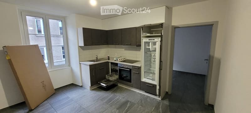 Modern kitchen with grey cabinets, white countertops, stainless steel appliances, and a large window. A cardboard box is visible on the left side.