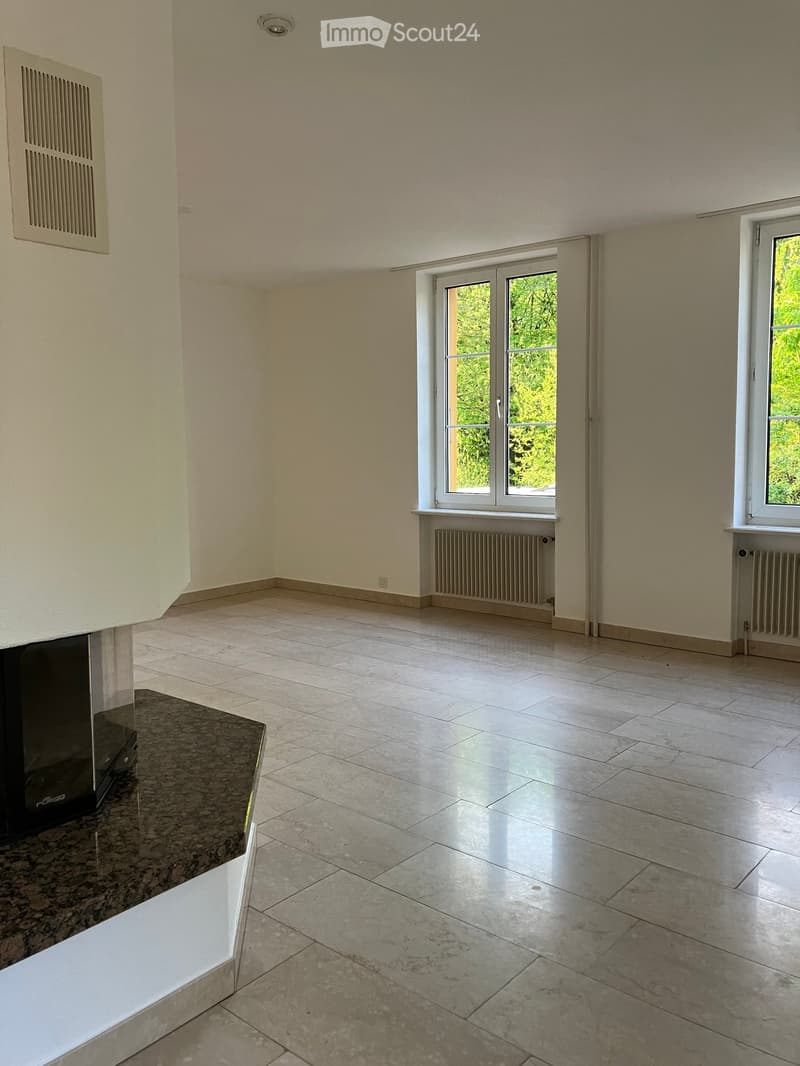 Empty room with light-colored walls, tiled floor, and large windows with white frames. Radiators are visible beneath the windows, and a small section of a dark countertop is visible in the foreground.