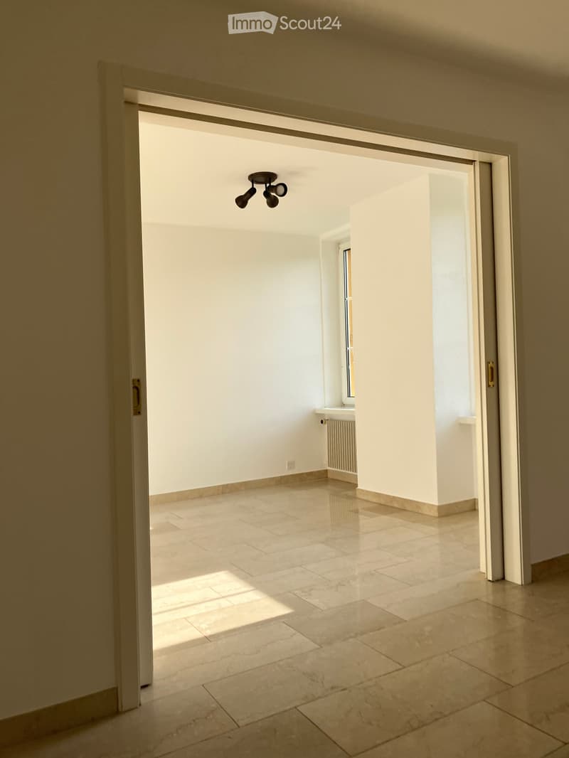 Empty room with beige tiled floor, white walls, and a small window letting in natural light. The room has a modern ceiling light fixture and a radiator near the window.