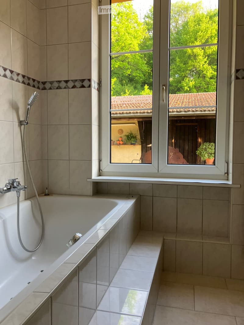 Bathroom with a bathtub, tiled walls, and a large window overlooking a garden area.