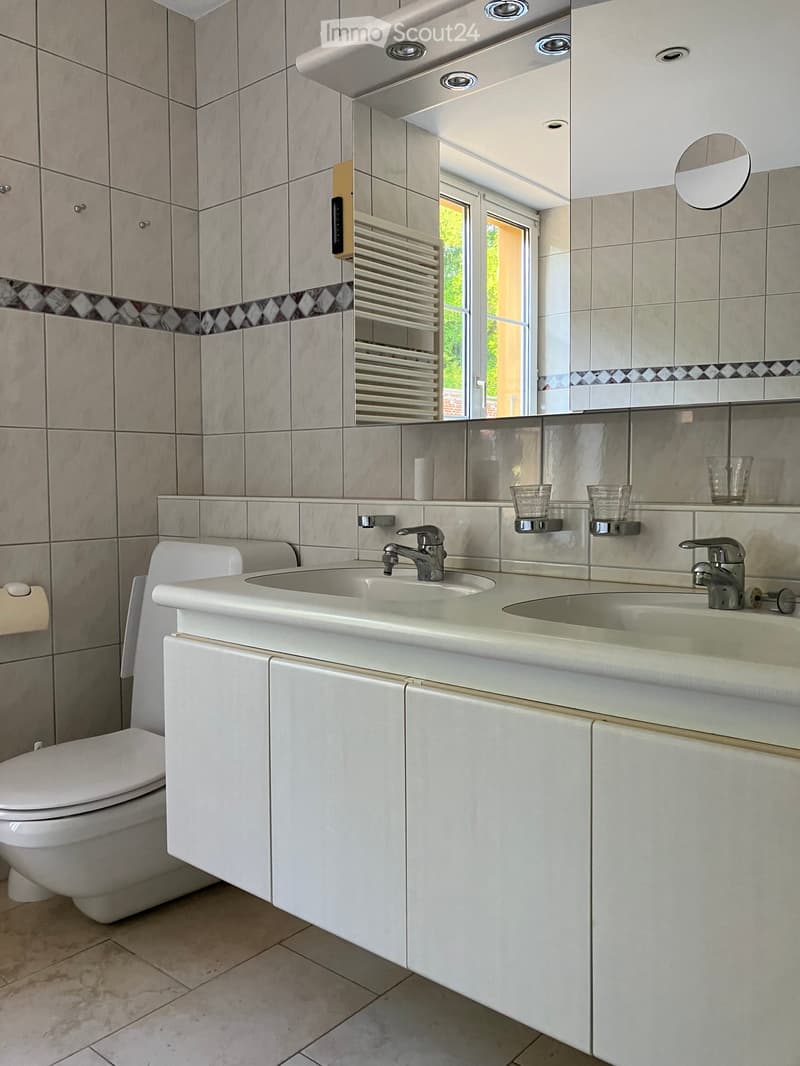 Modern bathroom with double sink, toilet, mirror, and radiator. Light gray tiles and white cabinetry.