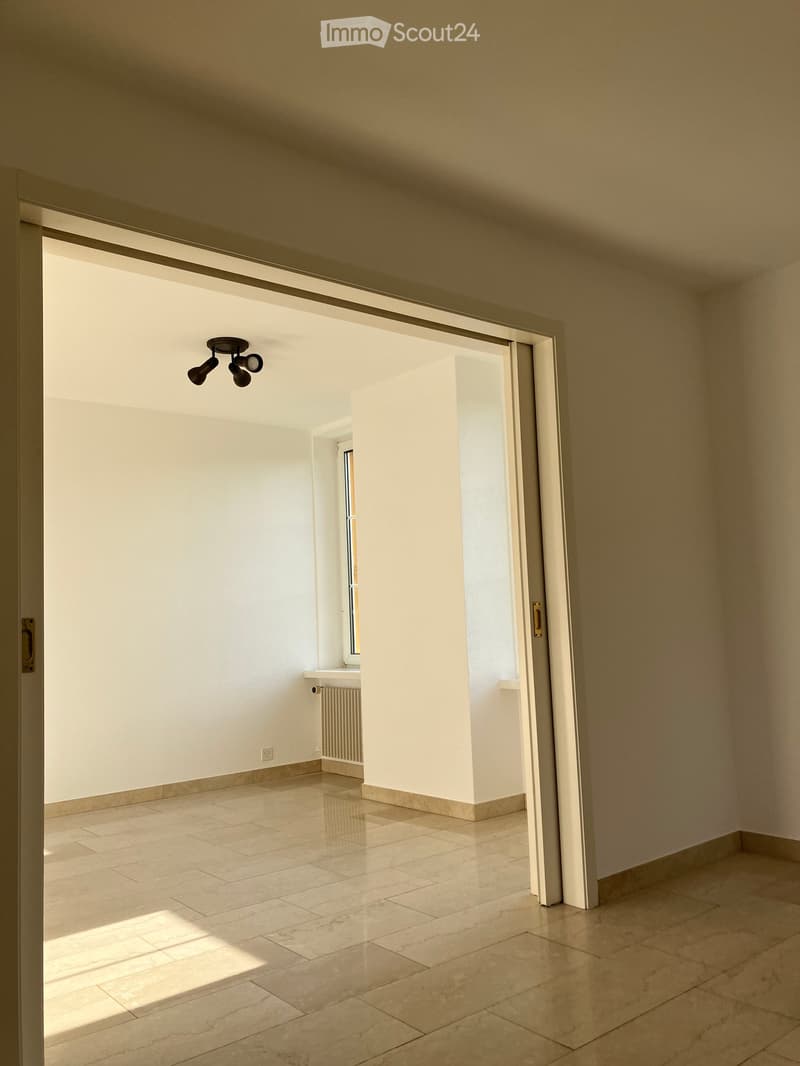 Empty room with light-colored tiled flooring, white walls, and a large window allowing natural light. Features a radiator and a ceiling light fixture.