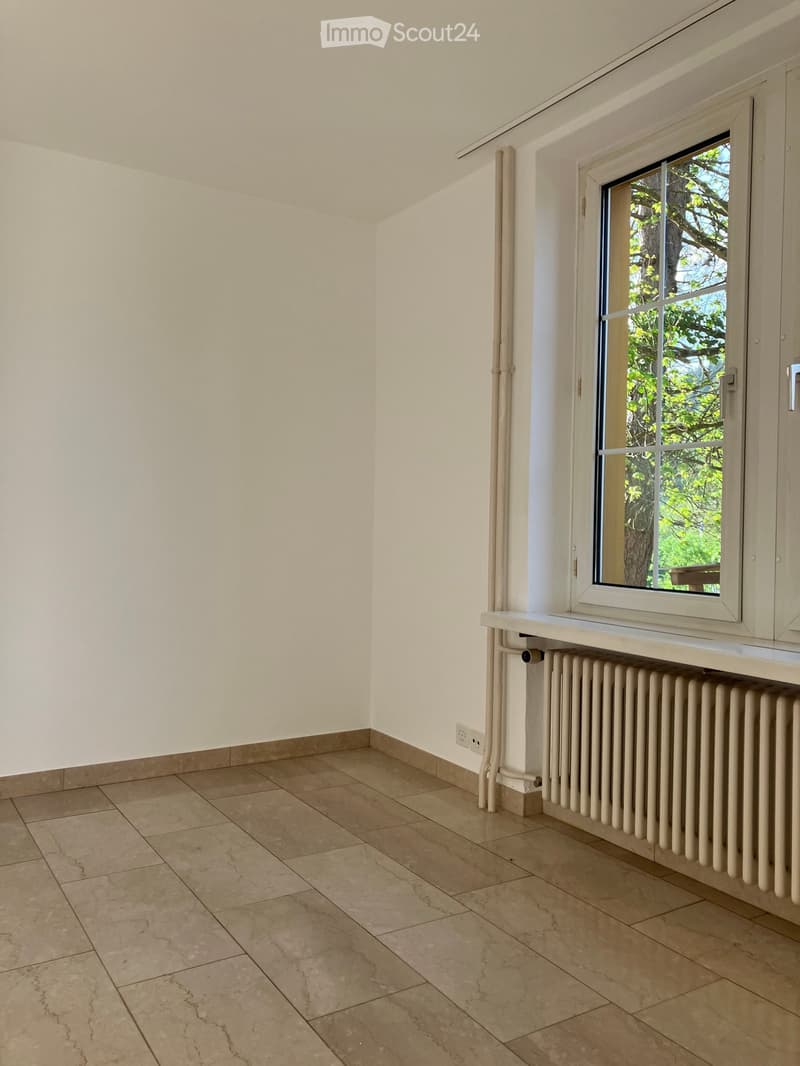 Empty room with beige tiled flooring, white walls, a radiator, and a window with a view of greenery outside.
