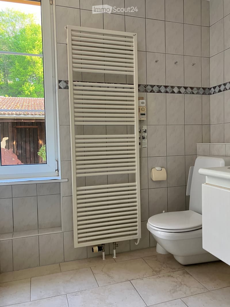 Bathroom with tiled walls, a toilet, a radiator, and a window with a view of greenery and a roofed structure.