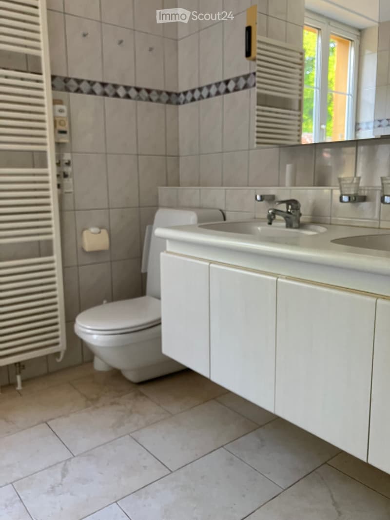 Modern bathroom with beige tiles, white sink, toilet, and radiator. Features a large mirror and a window with blinds.
