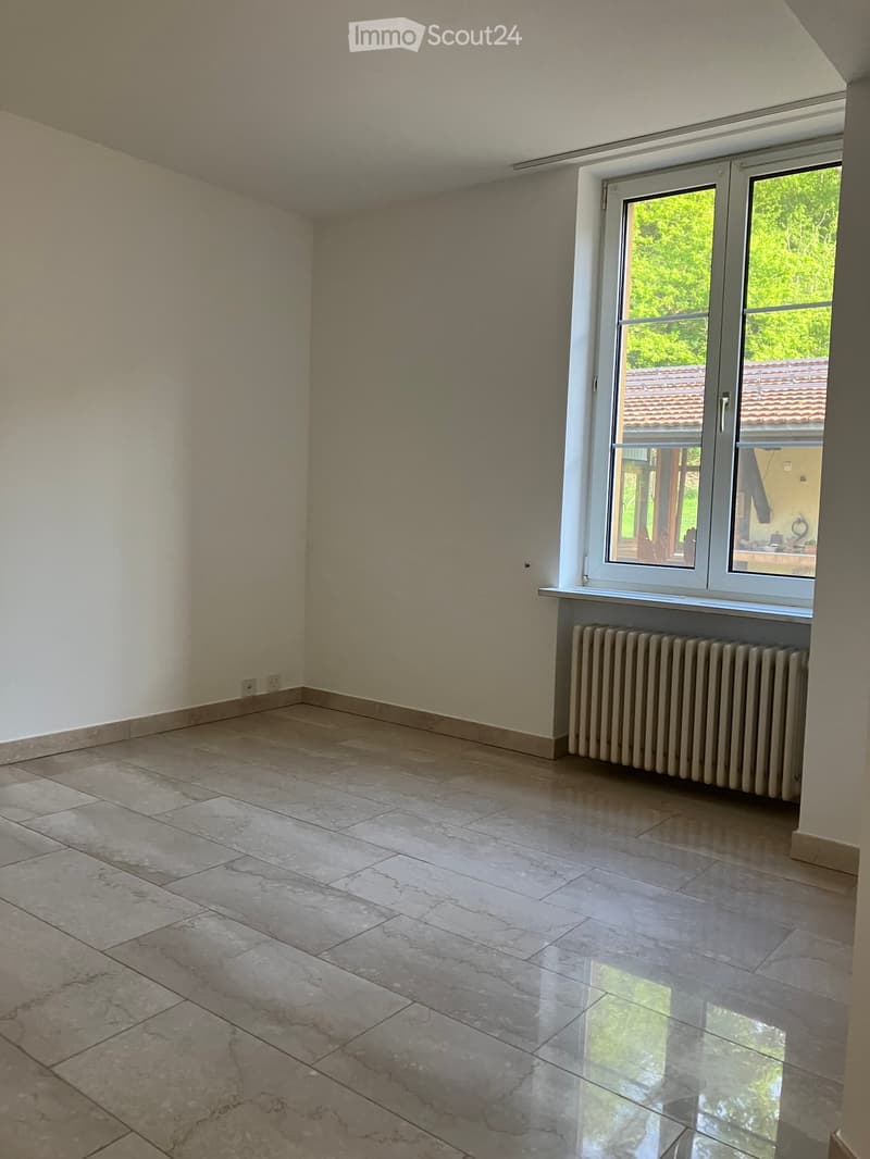 Empty room with tiled floor, white walls, large window with view of greenery, radiator under window, and visible electrical outlets on wall.