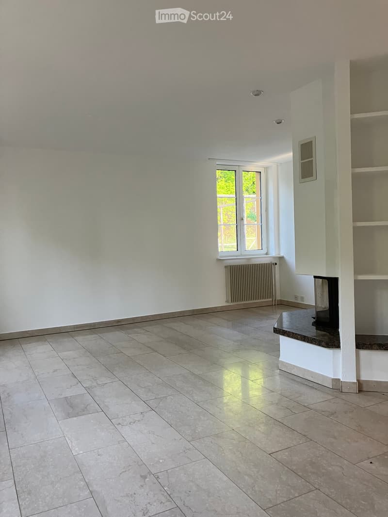 Empty room with light-colored tiled flooring, white walls, and a large window with white frames. A radiator is visible beneath the window, and a small built-in shelf unit is on the right side.