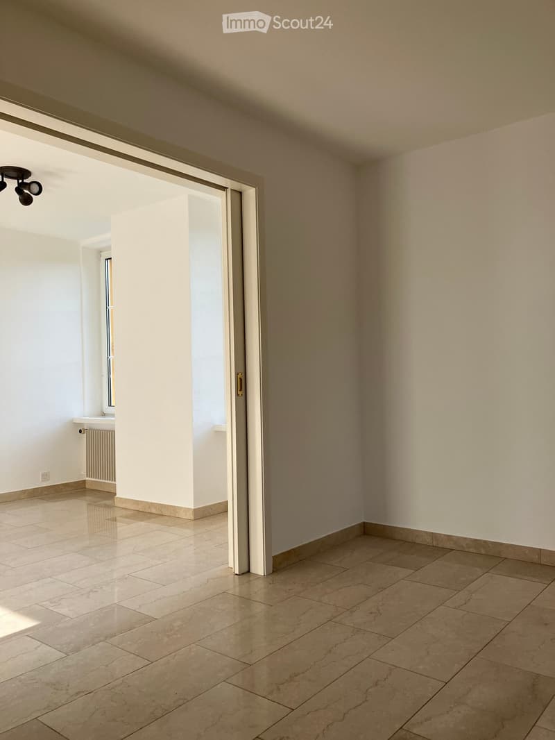 Empty room with tiled flooring, white walls, and a sliding door leading to another space. Natural light streams in from the adjacent room.