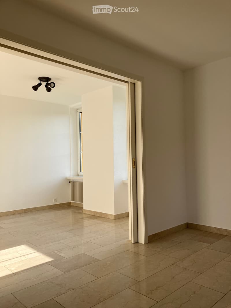 Empty room with beige tiled flooring, white walls, and a large open doorway leading to another space. A small window and radiator are visible in the adjacent area.
