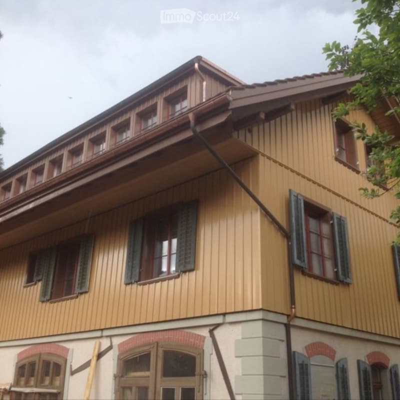 Two-story wooden building with a sloped roof, multiple windows with shutters, and a brick base.