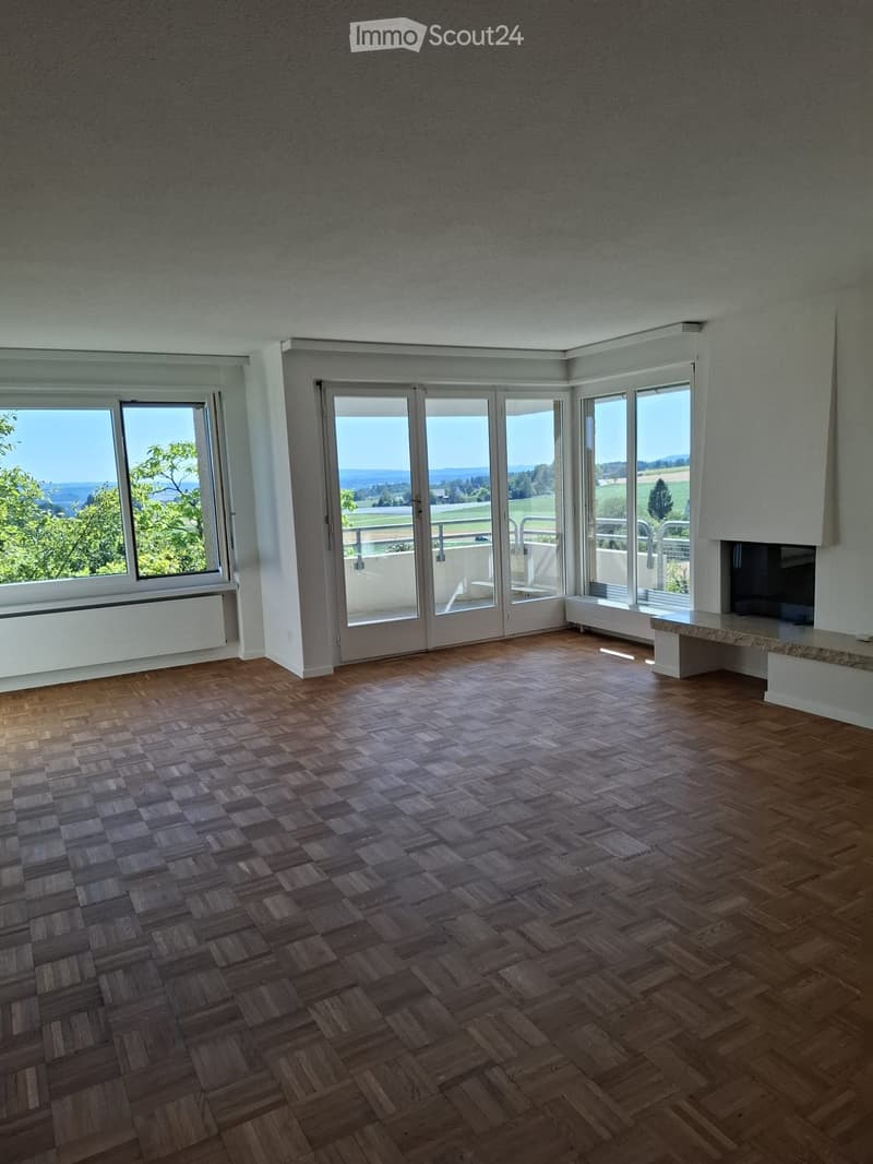 Empty room with wooden herringbone flooring, large windows and sliding glass doors leading to a balcony, offering scenic views of greenery and fields.