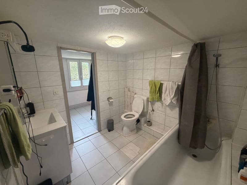 Bathroom with white tiled walls and floor, toilet, sink, mirror, and shower area. Towels are hanging on the wall, and a window provides natural light.