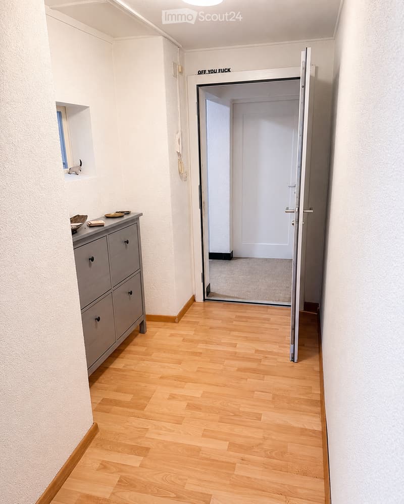 A narrow hallway with light wooden flooring and white walls, featuring a small cabinet with bowls and a partially open door leading to another room.