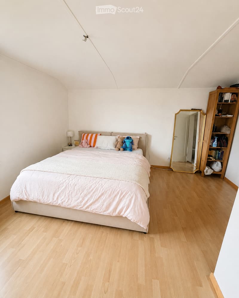 Cozy bedroom with light wooden flooring, a large bed with white bedding and colorful pillows, a nightstand with a lamp, a large mirror, and a wooden bookshelf filled with books and decorative items.