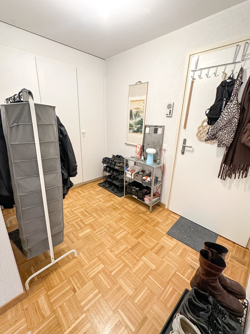 A well-lit hallway with wooden herringbone flooring, a shoe rack, a storage unit, a coat rack, a small table with a stool, and a mirror with a picture frame.