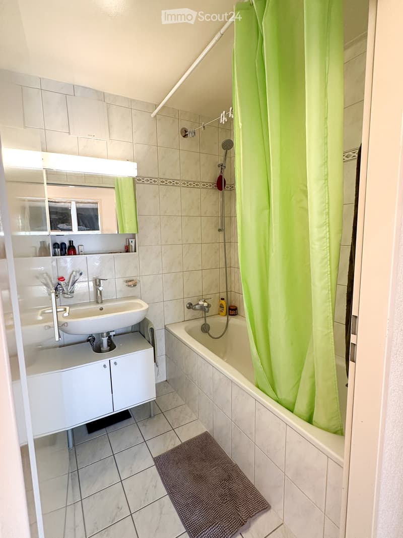 Modern bathroom with a white sink, bathtub, mirror, and tiled walls. Includes a green shower curtain, a small cabinet, and a gray bath mat.