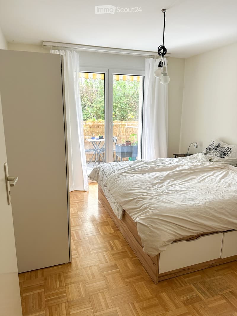 Bright bedroom with wooden flooring, large bed with white bedding, nightstand with lamp, and large windows with white curtains leading to a patio area with outdoor furniture.