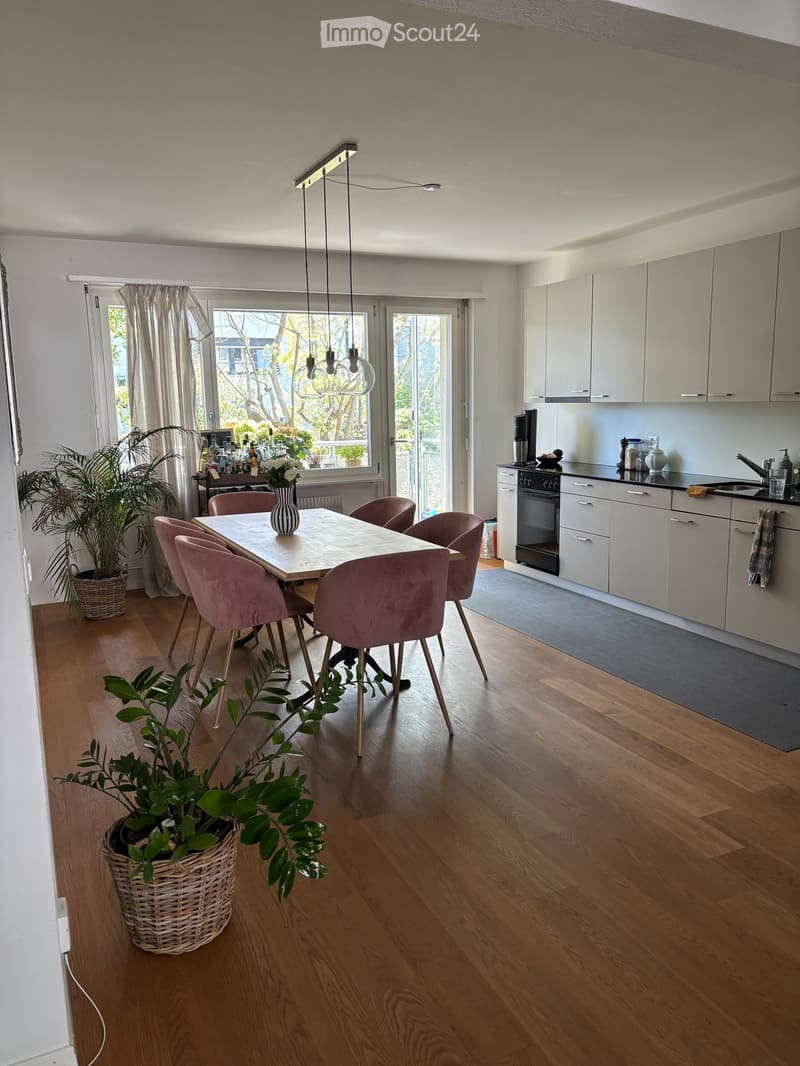 Modern kitchen with white cabinets, wooden flooring, and a dining area with a table and pink chairs. Features include pendant lights, a sink, and various kitchen appliances.