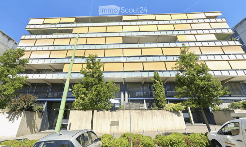Modern multi-story building with balconies and large windows, surrounded by greenery and parked cars.