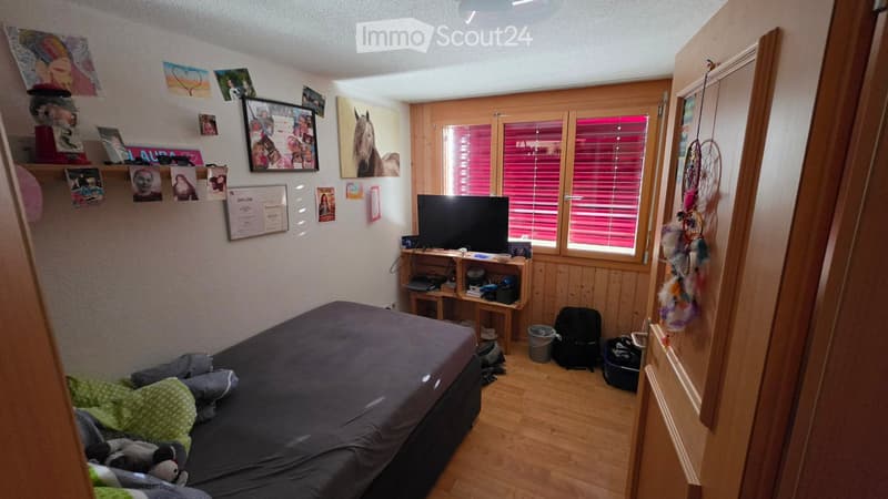 Cozy bedroom with wooden walls and flooring, featuring a bed, desk with a computer, and various decorations including picture frames and a dreamcatcher.