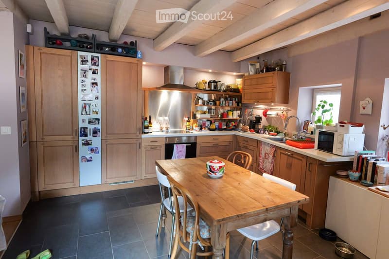 Bright kitchen with wooden cabinets, stainless steel appliances, a central dining table with chairs, and various kitchen items on counters and shelves.