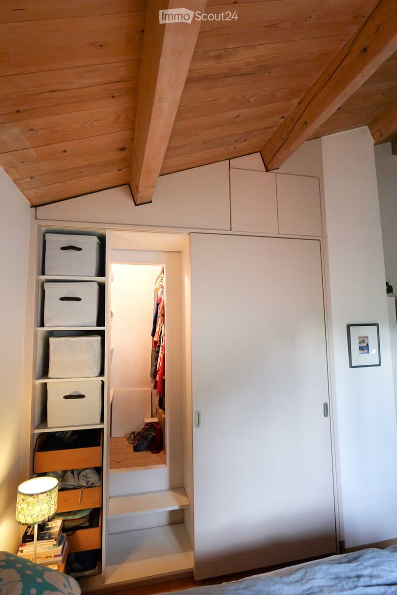 Cozy attic bedroom with wooden slanted ceilings, white built-in wardrobe, storage boxes, and a small lamp on a bedside table.