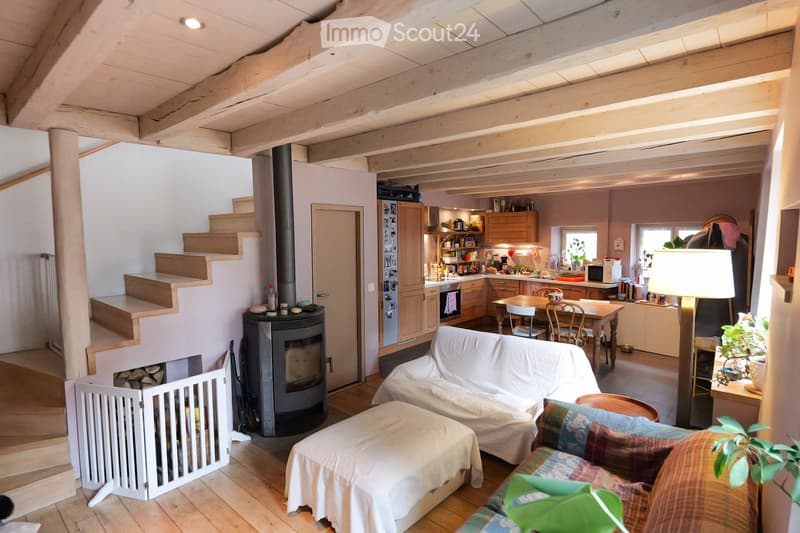 Open-plan living and dining area with exposed wooden beams, a covered sofa, ottoman, dining table with chairs, kitchen area with cabinets and appliances, and a wood-burning stove.