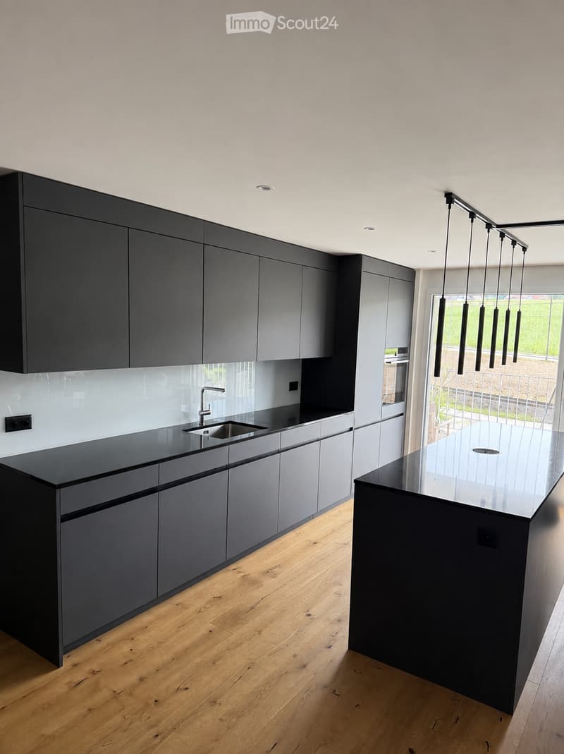 Modern kitchen with dark grey cabinets, a central island, and a sleek black countertop. Features a stainless steel sink and faucet, with pendant lights hanging above the island.