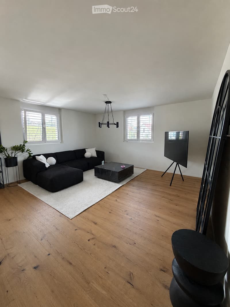 Modern living room with light wooden flooring, white walls, large windows, a black sectional sofa with white cushions, a black coffee table, a floor lamp, and a large screen on a stand.