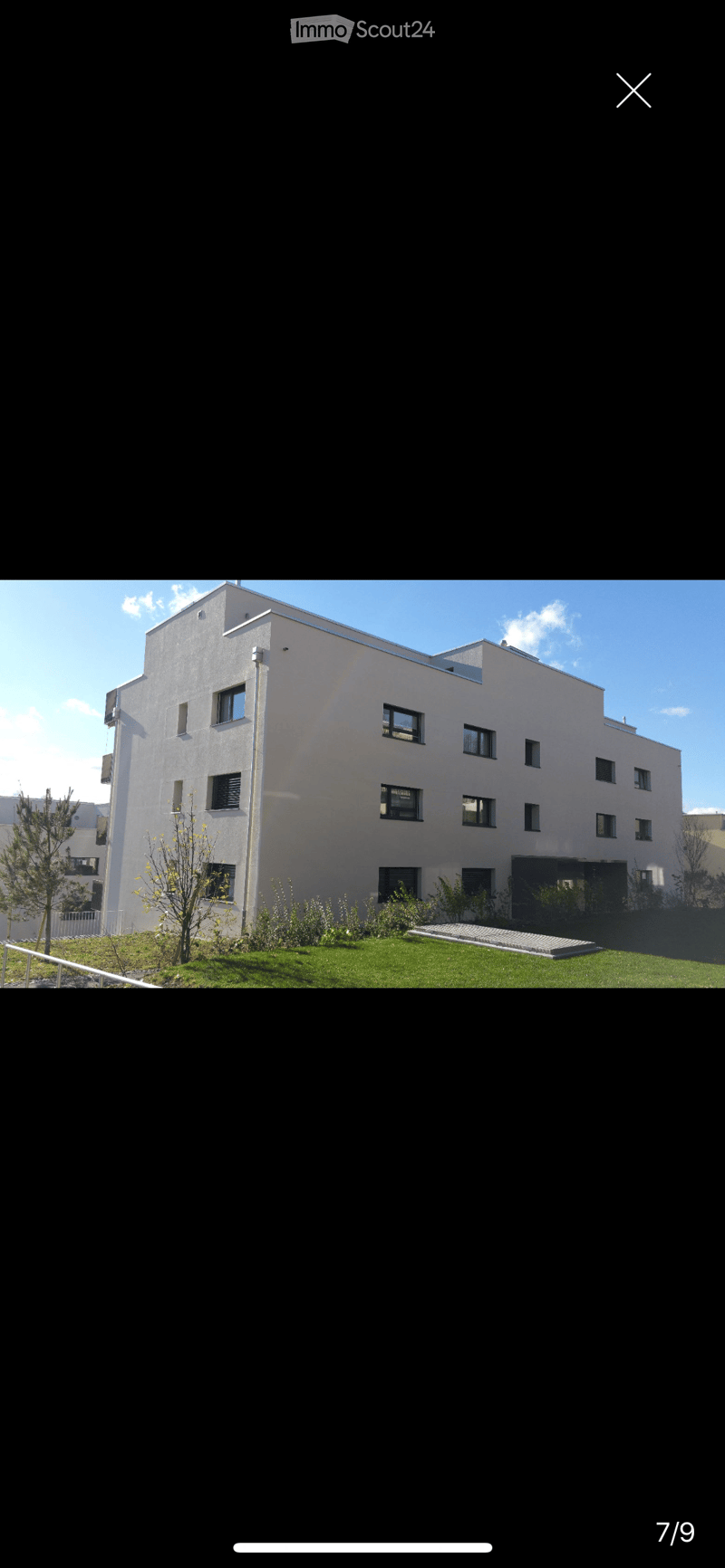 Modern multi-story building with white facade, multiple windows, and a small landscaped area in front.