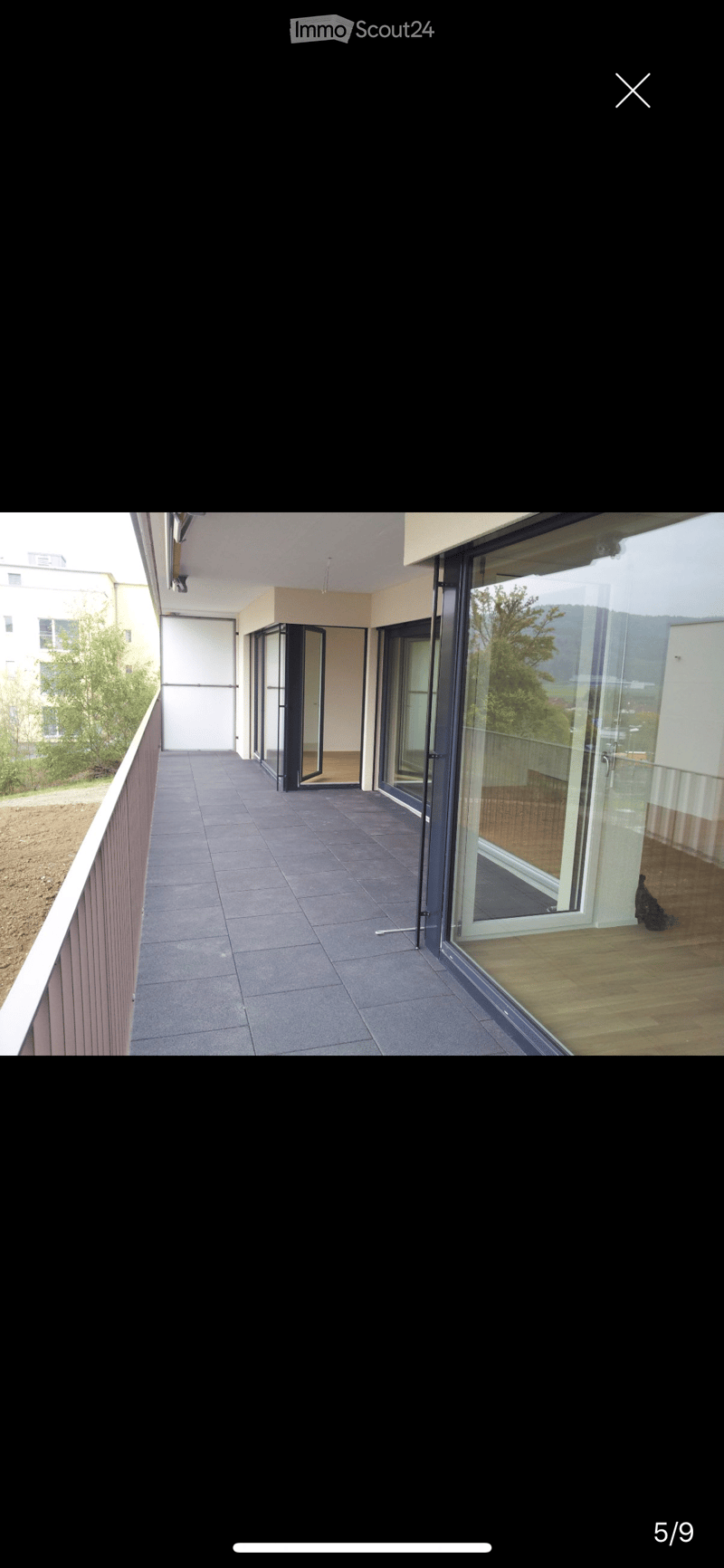 Modern balcony with grey tiled floor, glass sliding doors, wooden decking visible through the doors, and a view of greenery and buildings.