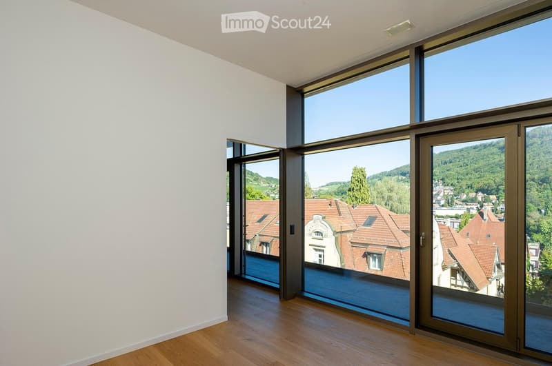 Empty room with large windows and wooden flooring, offering a scenic view of a town and surrounding hills.