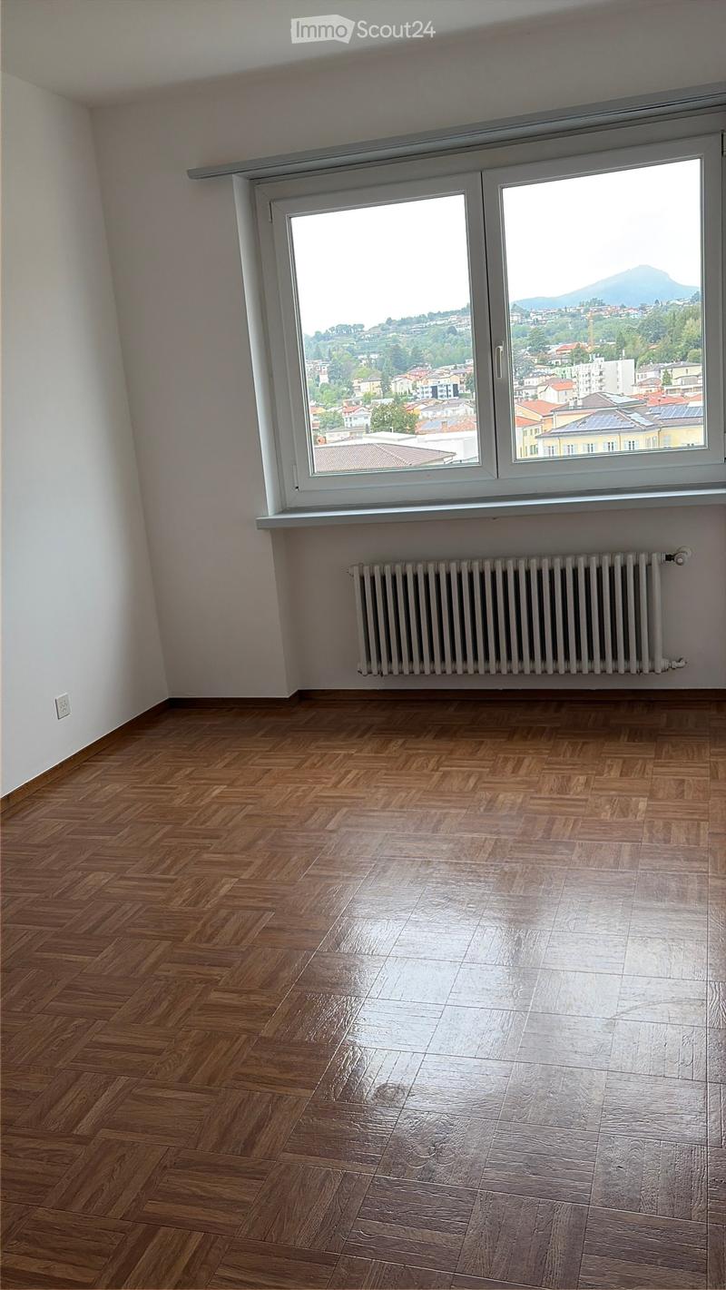 Empty room with wooden herringbone flooring, white walls, large window with cityscape view, radiator below window, and electrical outlet on wall.
