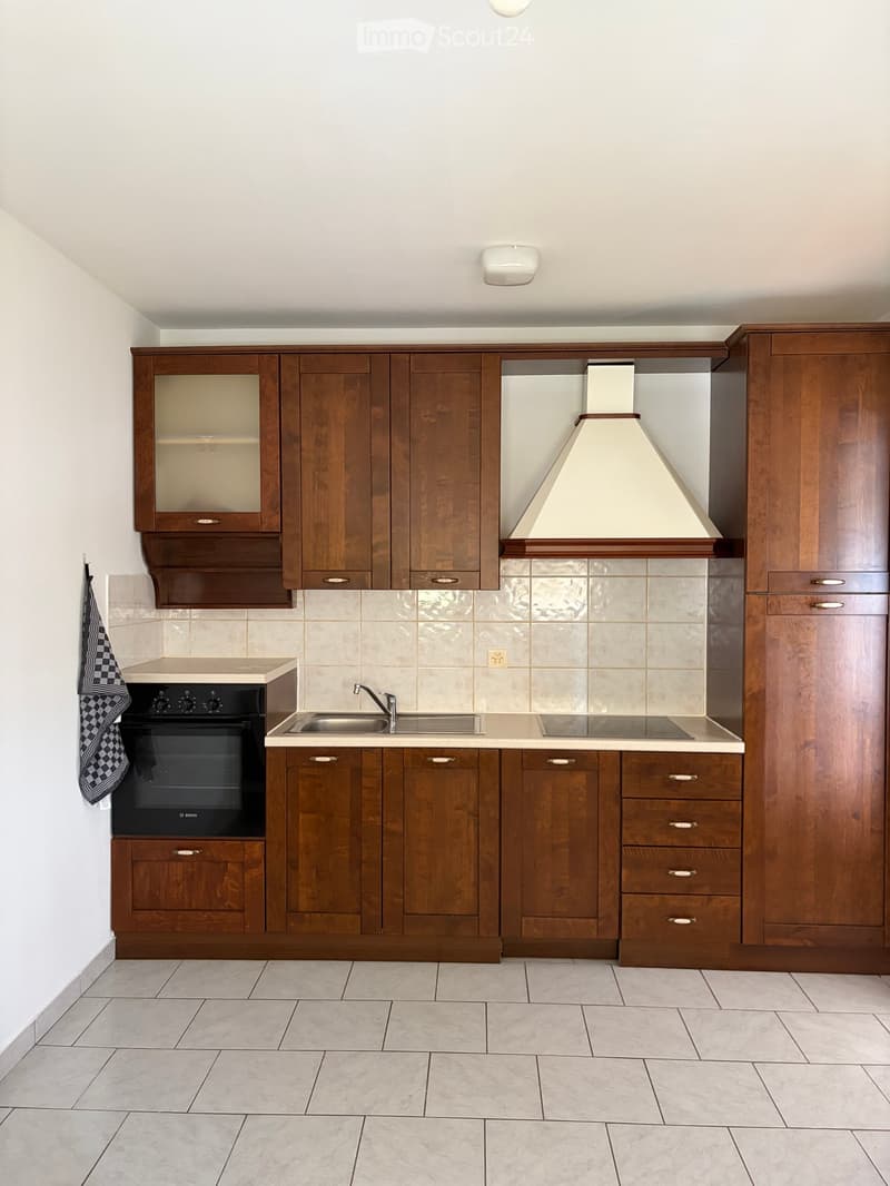 Modern kitchen with wooden cabinets, stainless steel sink, oven, and range hood. Light-colored tiles and a tiled floor.