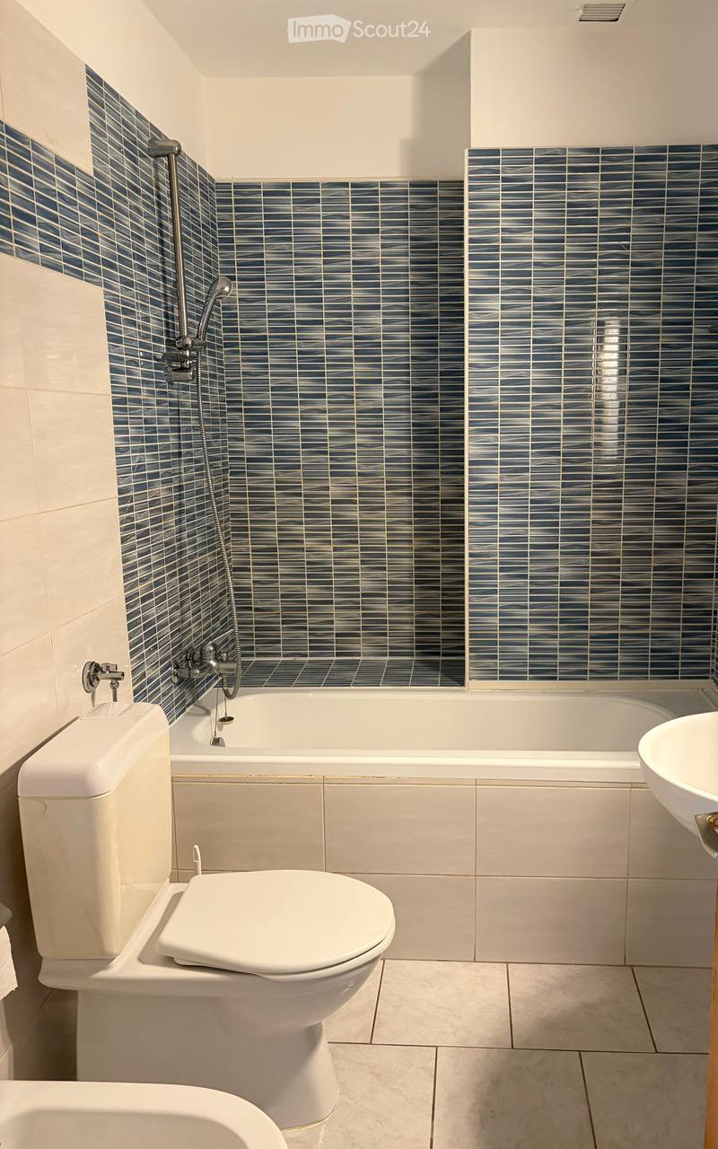 Modern bathroom with white tiles, a bathtub, toilet, and sink. Blue patterned tiles adorn the shower area.