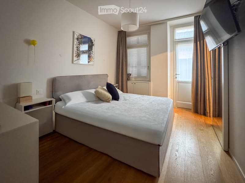 Modern bedroom with a large bed, nightstand, lamp, and artwork on the wall. The room features wooden flooring and large windows with curtains.