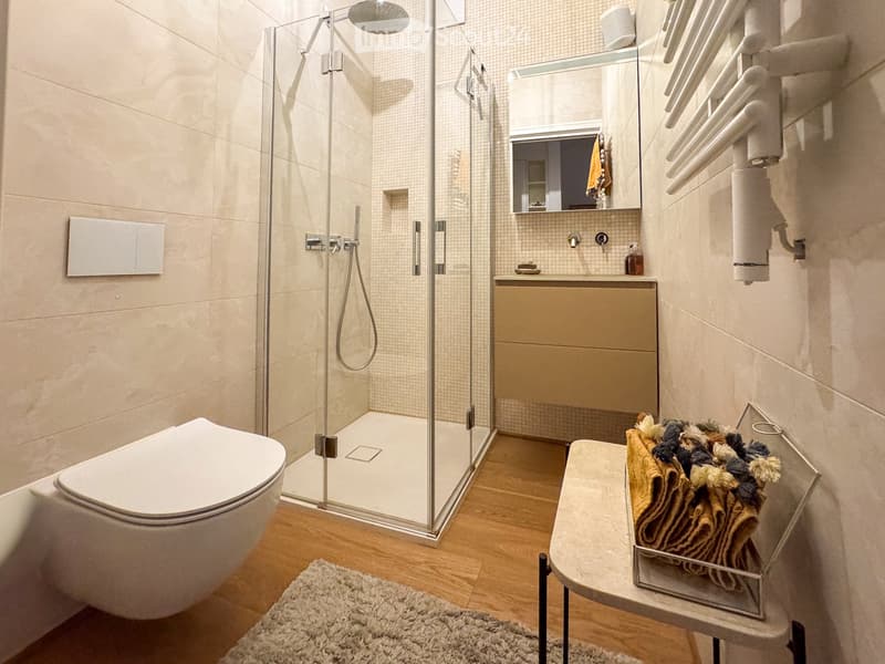 Modern bathroom with a glass-enclosed shower, toilet, sink, and towel rack. Beige tiles and wooden flooring.