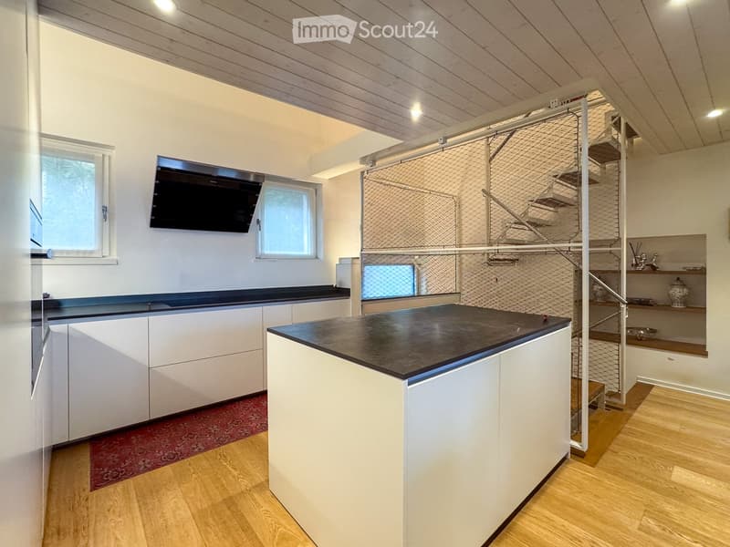 Modern kitchen with white cabinets, black countertops, wooden flooring, and a staircase leading to an upper level. A flat-screen screen is mounted on the wall.