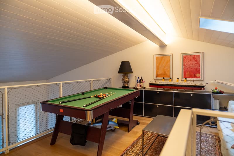 A well-lit room with a pool table, a table lamp, a model ship on a cabinet, framed artwork, and a small stool near the table.