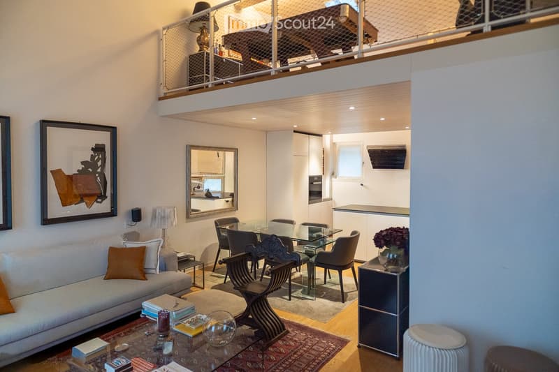 Modern living room with a sofa, coffee table, dining table, chairs, and a small kitchen area visible in the background.