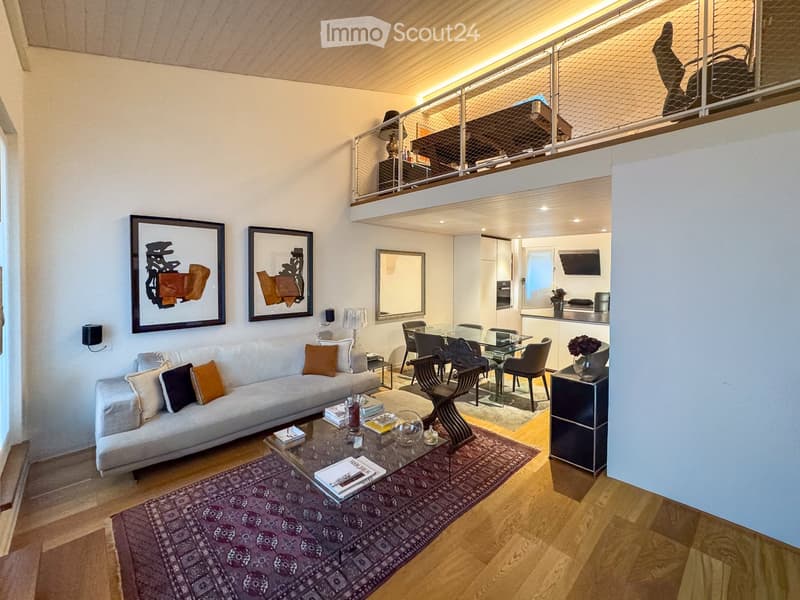 Modern living room with a grey sofa, coffee table, dining area, and artwork on the walls. The room features wooden flooring and a lofted balcony area.