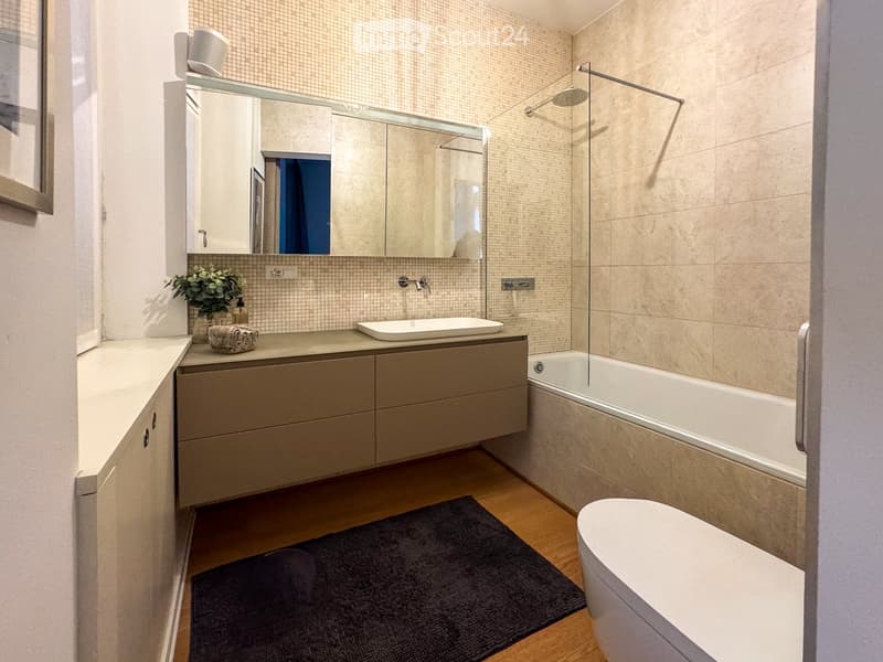 Modern bathroom with beige tiles, a large mirror, a sink with a cabinet, a bathtub, a toilet, and a small plant on the counter.