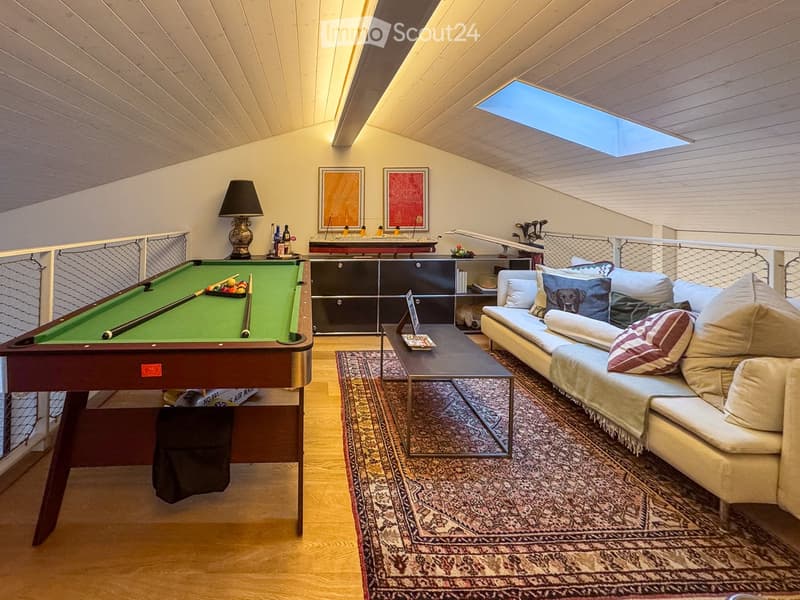 A cozy attic room with wooden floors, a pool table, a sofa, a coffee table, a table lamp, and framed artwork on the walls.