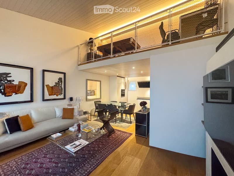Modern living room with a white sofa, coffee table, dining area, and artwork on the walls. The room features wooden flooring and a lofted area visible in the background.