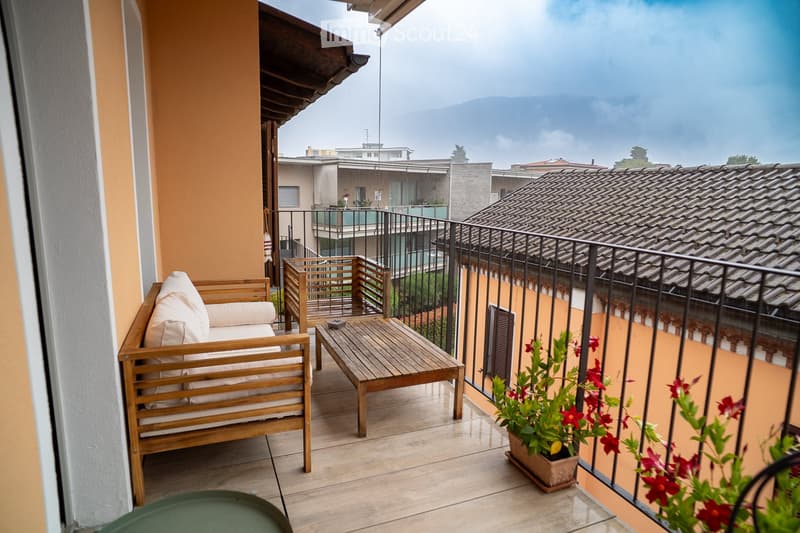 Balcony with wooden sofa, small table, and potted plants, overlooking a cityscape with tiled roofs and distant mountains.